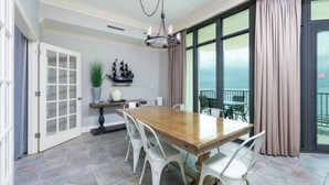 A dining area with a wooden table and white chairs, a decorative ship model, and a view of the ocean through large windows.