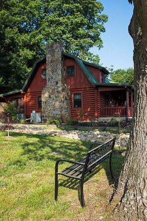 A log cabin with a stone chimney, surrounded by a grassy area and a bench. - Cozy Cabin, Leaves 🍁 Private Swim Jacuzzi Spa- Beautiful Gardens & Orchard   (Jonesborough)