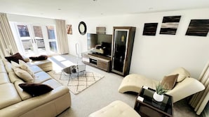 A modern living room with a sofa, ottoman, and a coffee table. There is a television and a cabinet in the room. - Horley Hideaway - 4 Bedroom House- Sleeps 7 (Horley)