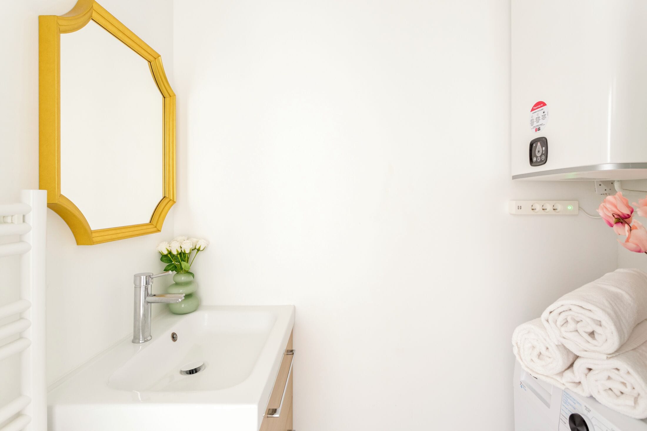 A bathroom with a white sink, a yellow-framed mirror, a green vase with white flowers, and a stack of white towels.