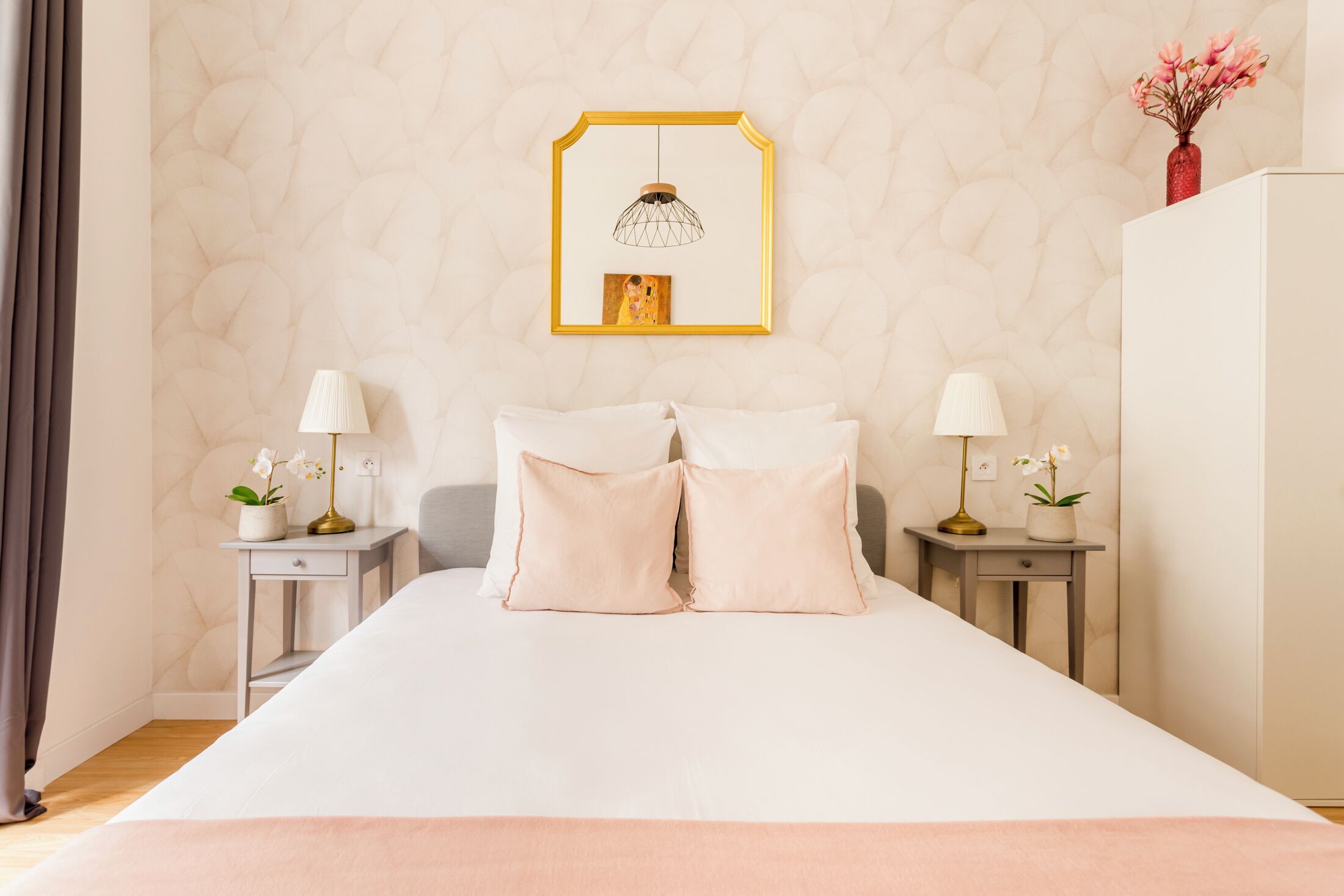 A neatly arranged bedroom with a large bed, two bedside tables with lamps, a framed artwork above the bed, and a vase with flowers on the dresser.