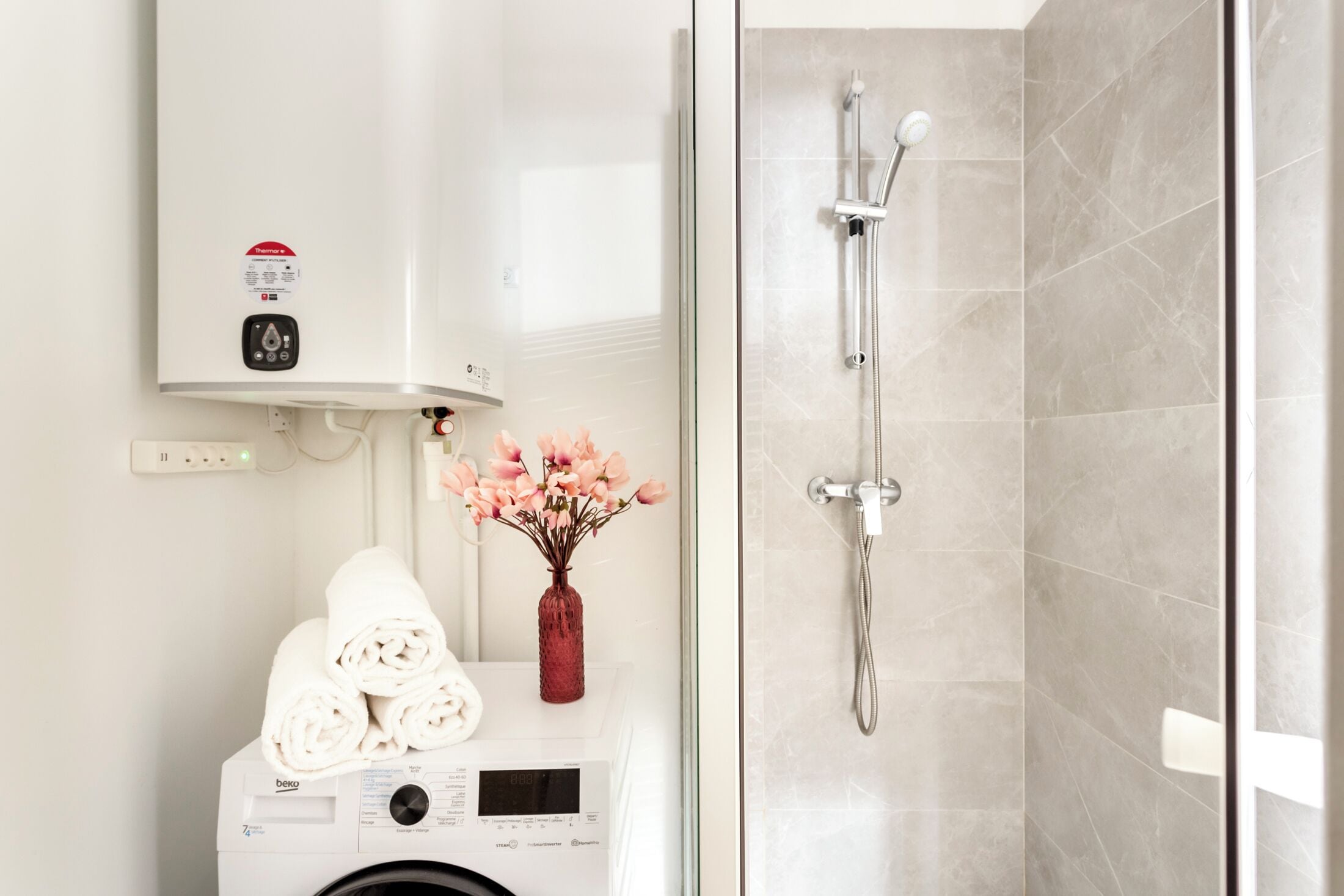 A bathroom with a wall-mounted heater, a glass-enclosed shower, a washing machine, and a vase with pink flowers.
