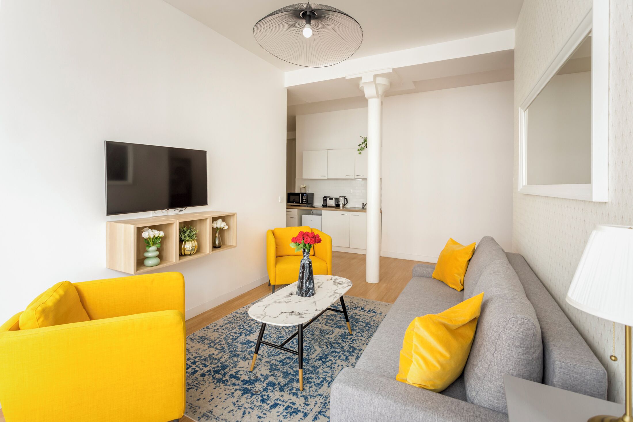 Un salon moderne avec un canapé gris, des fauteuils jaunes, un téléviseur à écran plat et une petite table basse sur laquelle est posé un vase de fleurs.