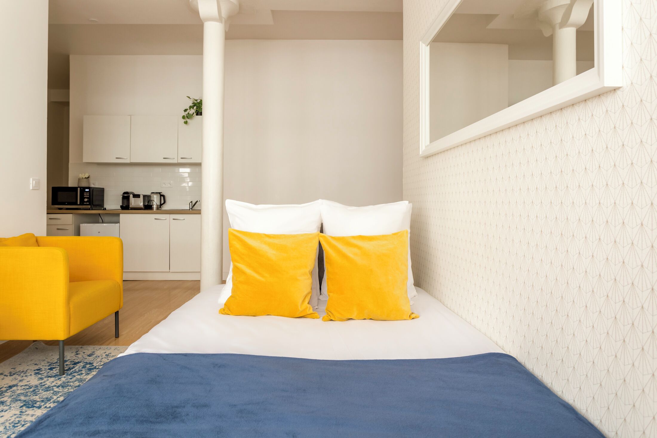 A bedroom with a bed, two yellow pillows, a blue blanket, a yellow chair, and a view of a kitchen in the background.