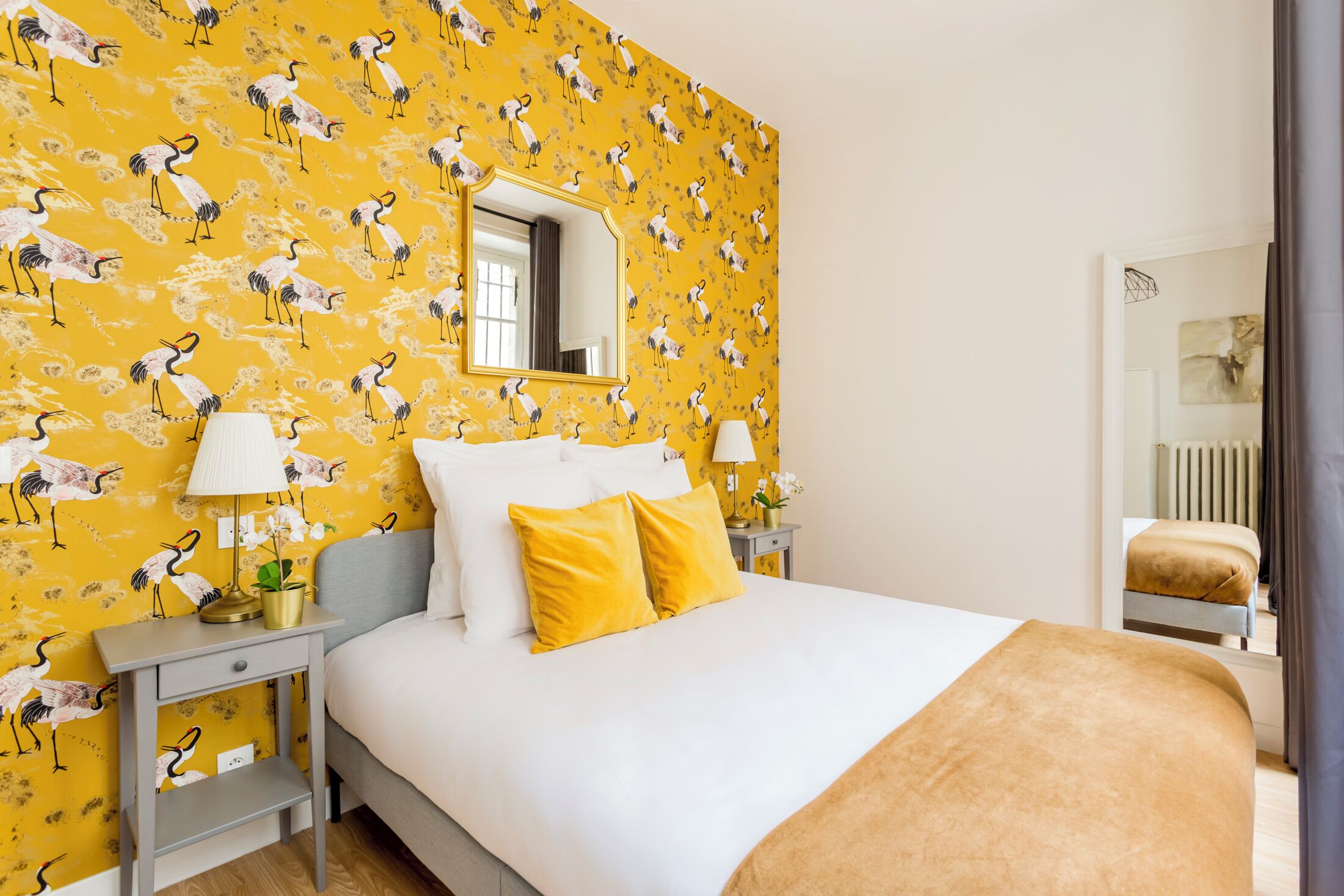 A hotel room with a bed, two bedside tables, a mirror, and a yellow patterned wallpaper.
