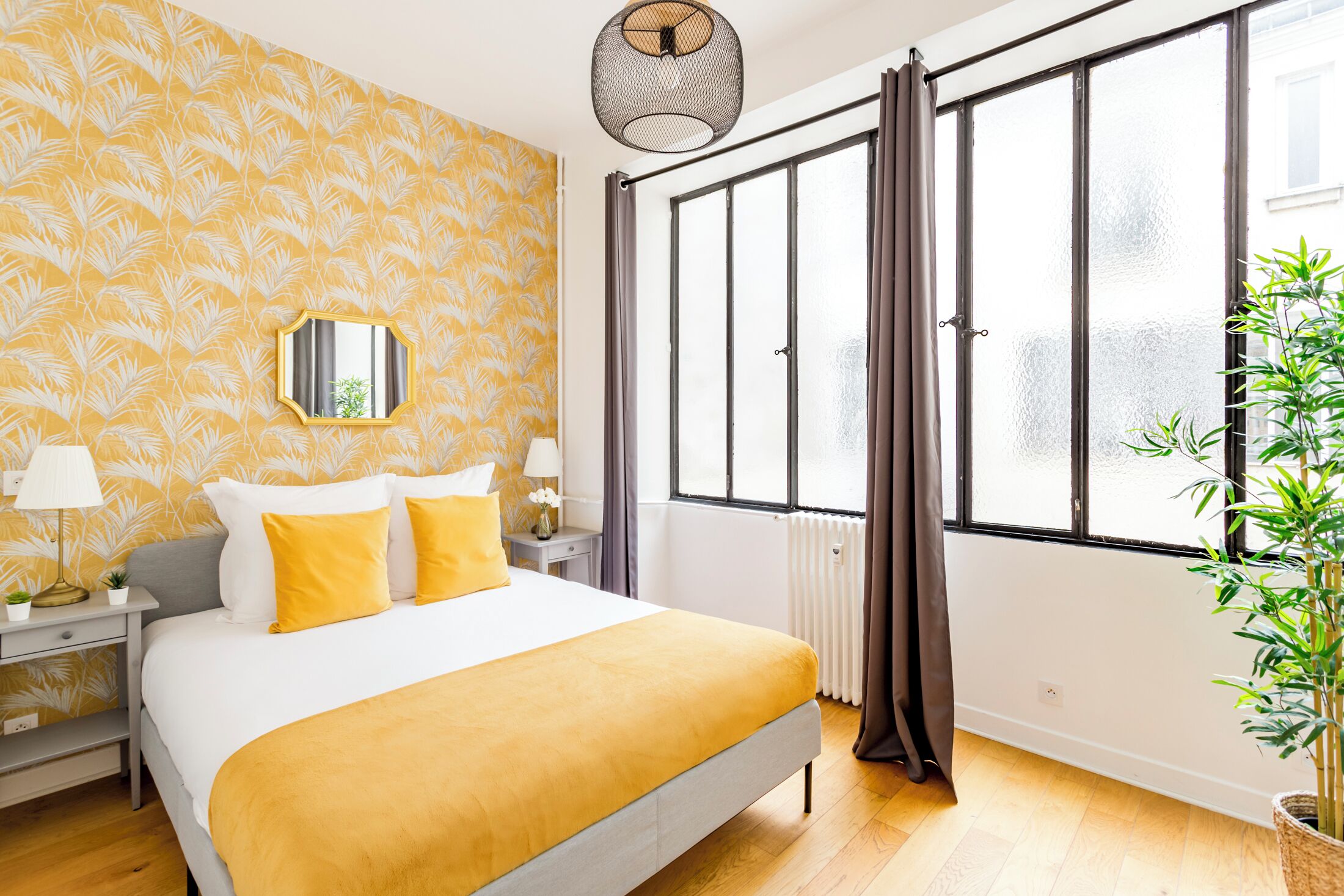 Une chambre d’hôtel avec un grand lit, une table de chevet, un miroir, une fenêtre avec des rideaux et une plante en pot.