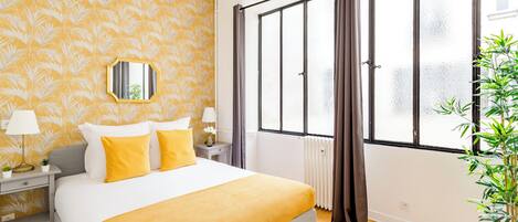 Une chambre d’hôtel avec un grand lit, une table de chevet, un miroir, une fenêtre avec des rideaux et une plante en pot.