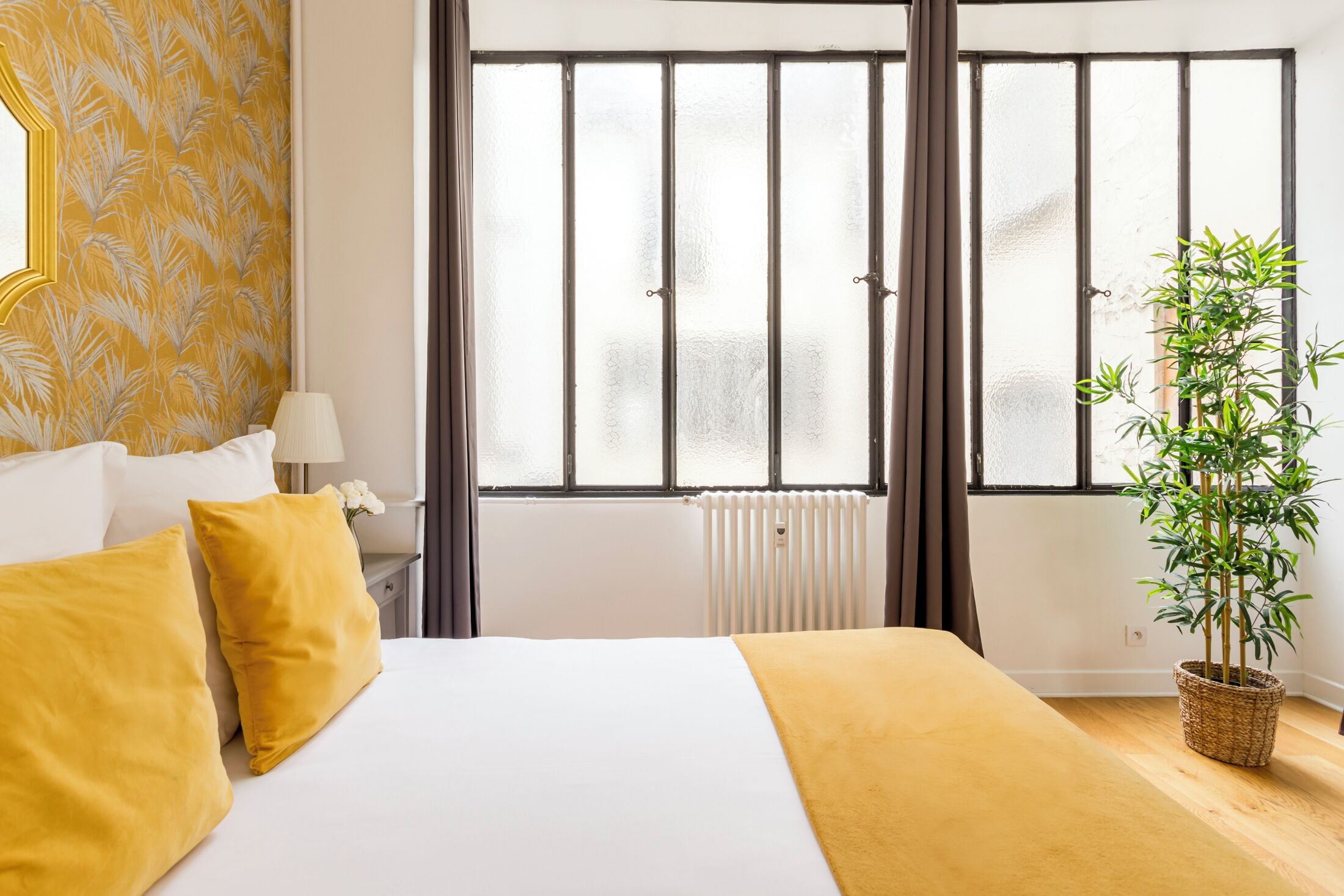 Habitación de hotel con cama, ventana con persianas, radiador, mesita de noche con lámpara y una planta en maceta.