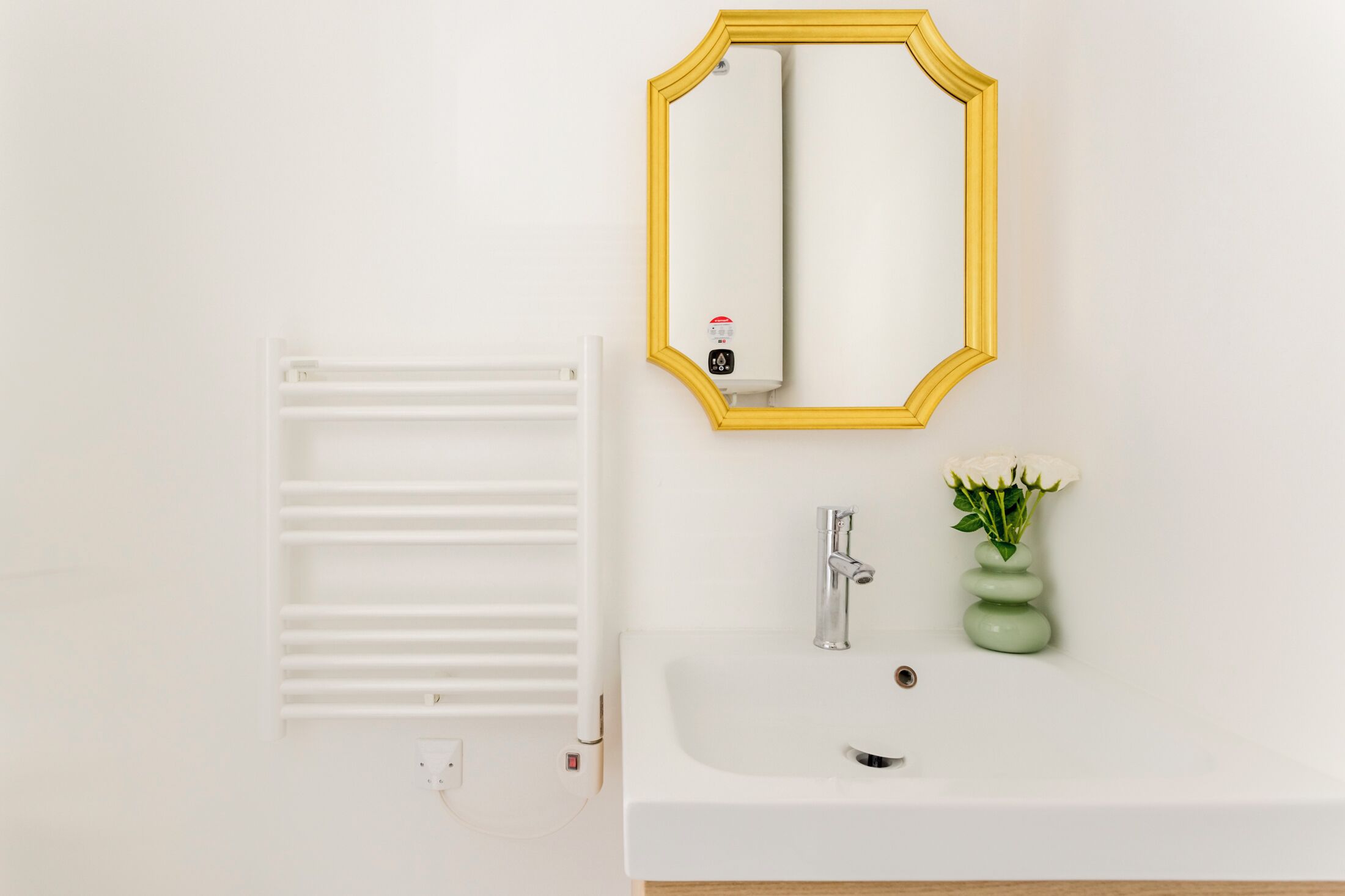 Baño con lavamanos blanco, espejo con marco amarillo, radiador blanco y un jarrón verde con flores blancas.