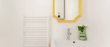 Un bagno con un lavandino bianco, uno specchio con cornice gialla, un termosifone bianco e un vaso verde con fiori bianchi.