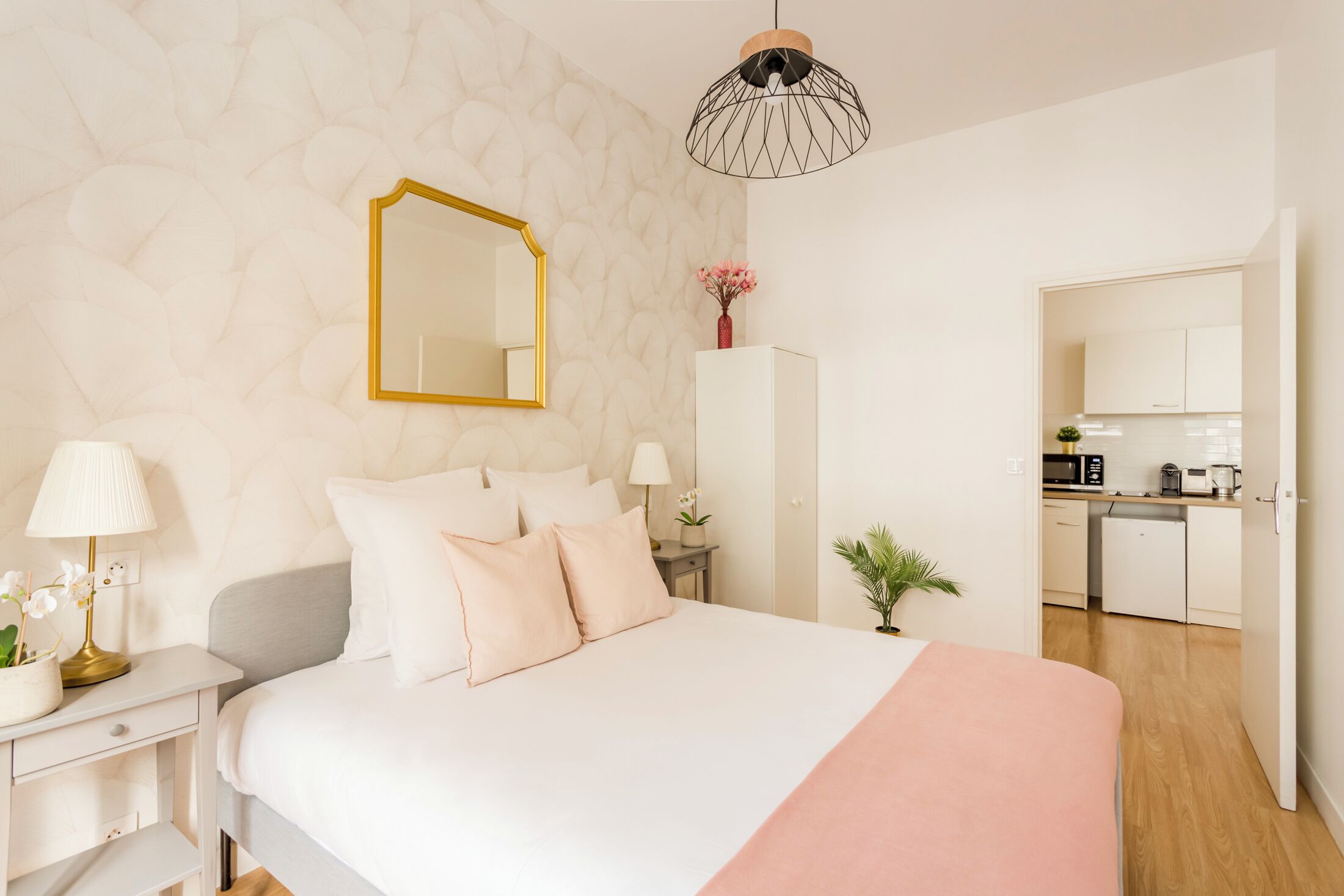 Ein Schlafzimmer mit Bett, Nachttischen, einem Spiegel und einer kleinen Küchenzeile im Hintergrund.
