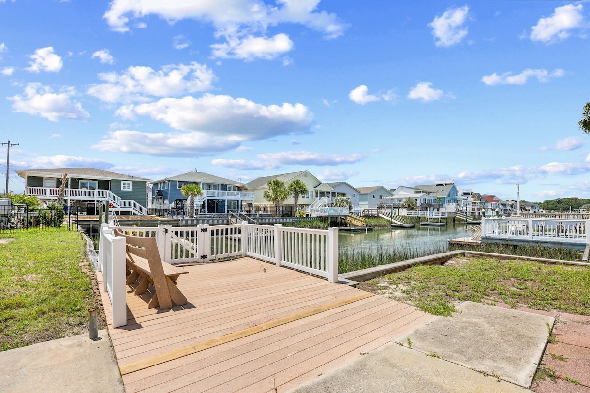 A waterfront property with a wooden dock, a marina with boats, and houses in the background.