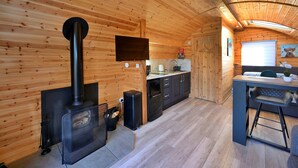 A cozy cabin interior with a wood-paneled ceiling, a central fireplace, a kitchen area with a sink and countertop, and a dining space with a table and chairs.