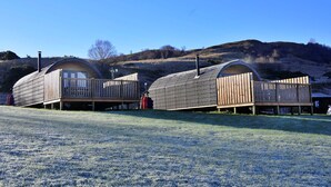 Two wooden cabins with curved roofs and wooden decks, situated on a grassy field with a hilly backdrop.