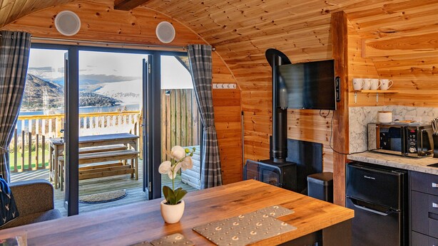 Innenansicht einer Holzhütte mit Küche, Essbereich und großem Fenster mit Blick auf eine reizvolle Landschaft.