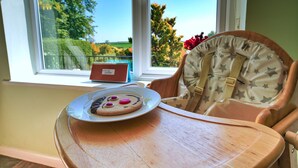 A high chair with a star-patterned cushion and a plate with cookies and a red object on a wooden table near a window with a view of trees and a fence. - Bryn Tor (Bolsover)