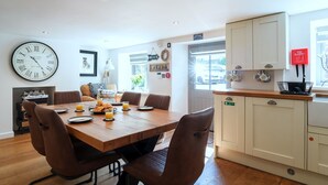 Un comedor con una mesa de madera y sillas marrones, un reloj en la pared, y una cocina con armarios blancos y un letrero rojo.