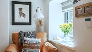 Una habitación con un sillón de cuero, una ilustración enmarcada de un perro, una lámpara blanca y una ventana con flores.
