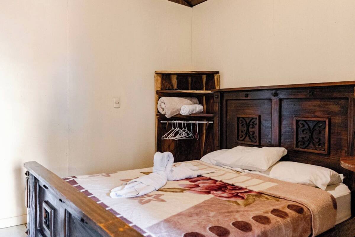 A neatly made bed with a floral-patterned bedspread, a wooden headboard with decorative carvings, and a small wooden shelf with towels.