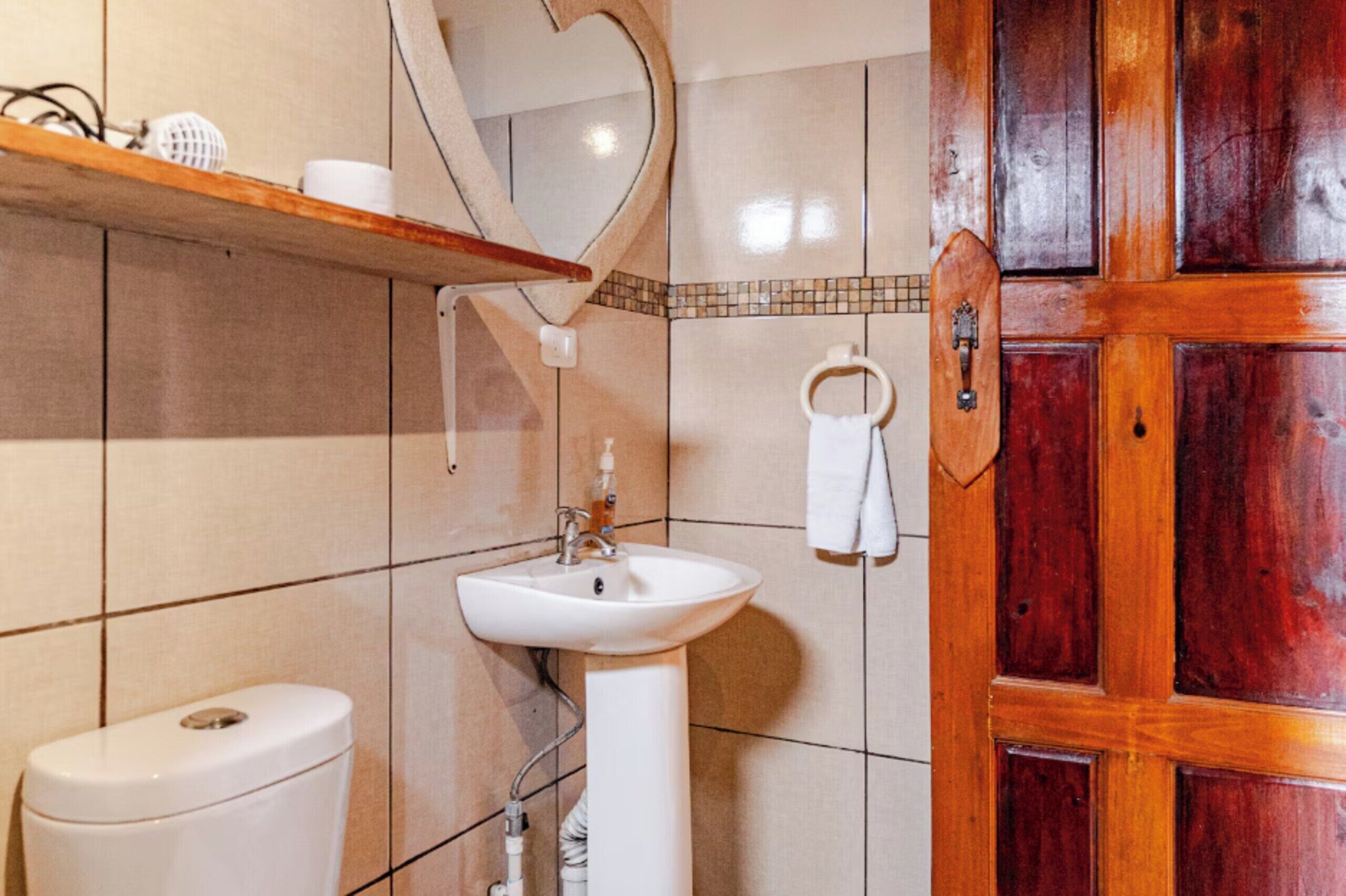 Un baño con un lavabo blanco, un inodoro, un estante de madera con artículos de aseo, un espejo y una puerta de madera.
