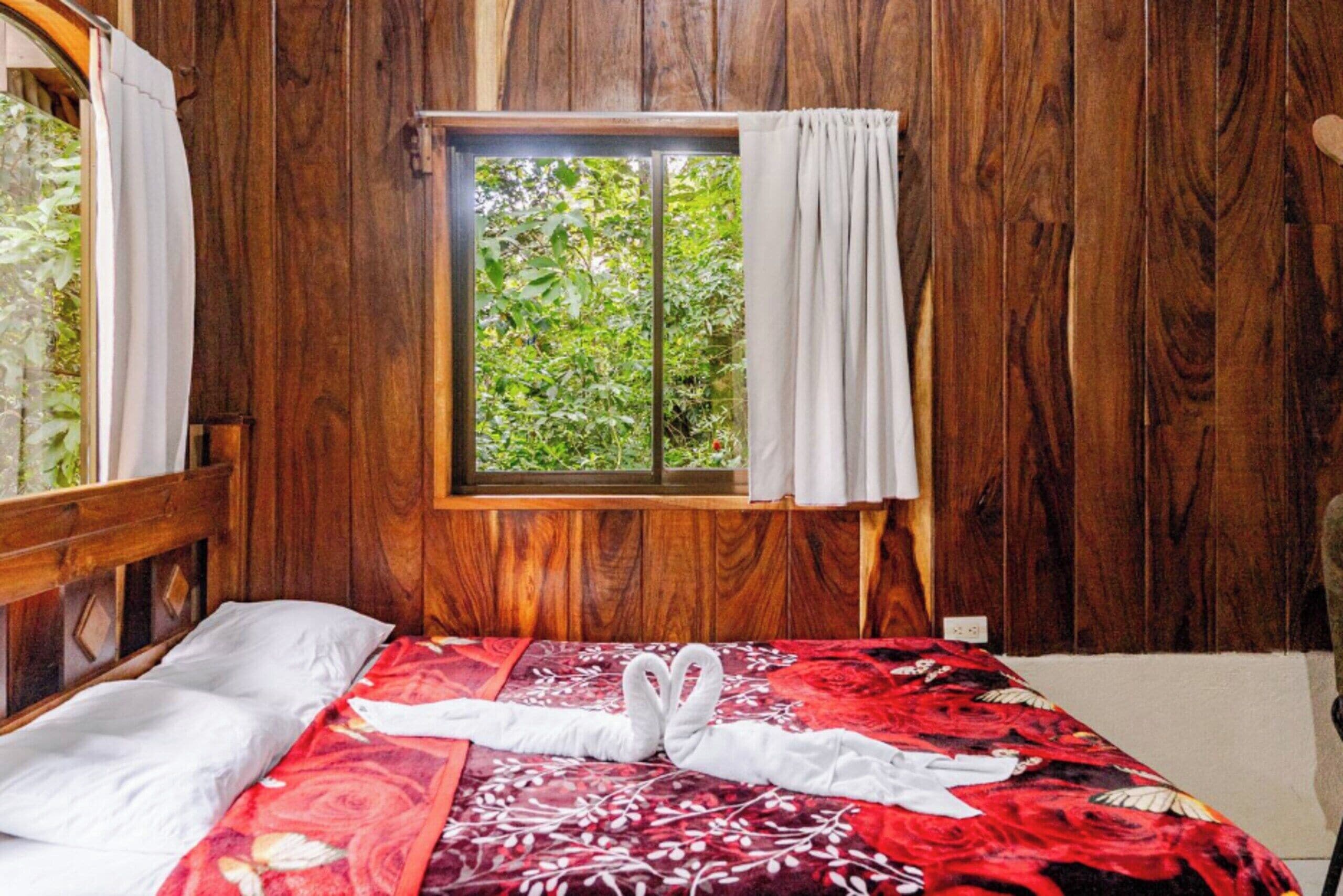 A wooden-paneled room with a bed, a window with curtains, and a view of greenery outside.