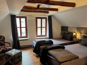 Een slaapkamer met een schuine plafond, twee ramen met gordijnen, een bed met kussens, een nachtkastje met een lamp en een fauteuil met een kussen.