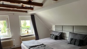 A bedroom with a large bed, wooden beams on the ceiling, two windows with curtains, and a radiator.