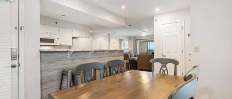 A dining area with a wooden table and chairs, white cabinets, and a kitchen in the background.
