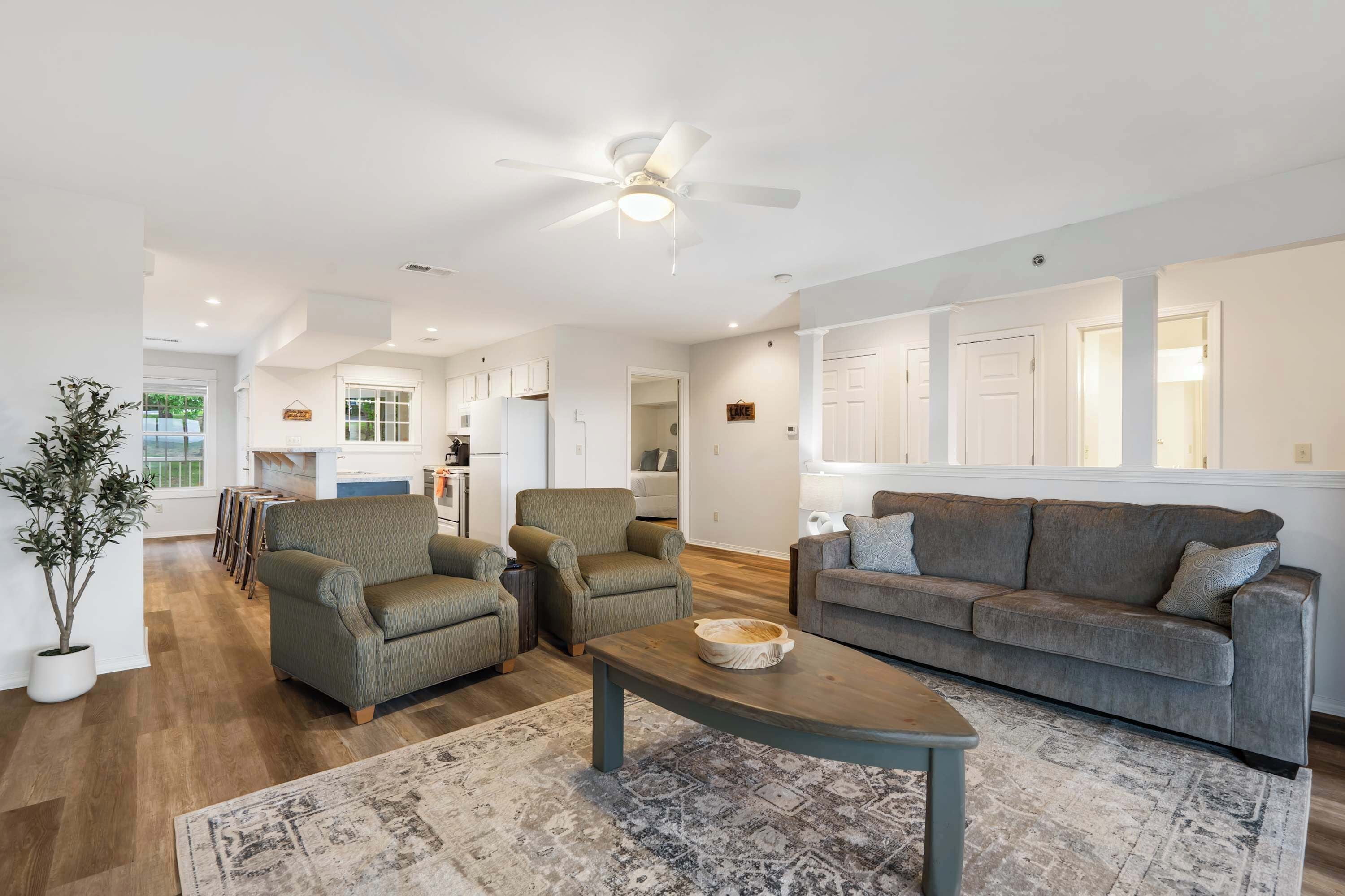 A spacious living room with a grey sofa, two armchairs, a coffee table, and a large window.