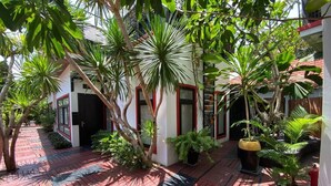A courtyard with a red brick pathway, surrounded by lush greenery and a building with white walls and red trim.
