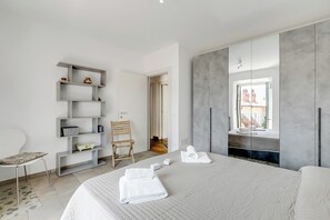 A modern hotel room with a large bed, a built-in wardrobe, a seating area with a chair, and a view of the outside.