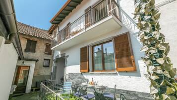 A two-story house with a balcony, a dining area with outdoor furniture, and a garden.