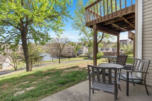 A covered patio with outdoor furniture, overlooking a grassy area and a body of water with buildings in the background. - Pond View Serenity For 6! (Branson)