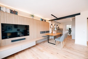 A modern living room with a large flat-screen TV, wooden panel wall, dining table with chairs, and a kitchen in the background. - Apartment 'Tauberblick' with Shared Terrace, Private Garden and Wi-Fi (Röttingen)