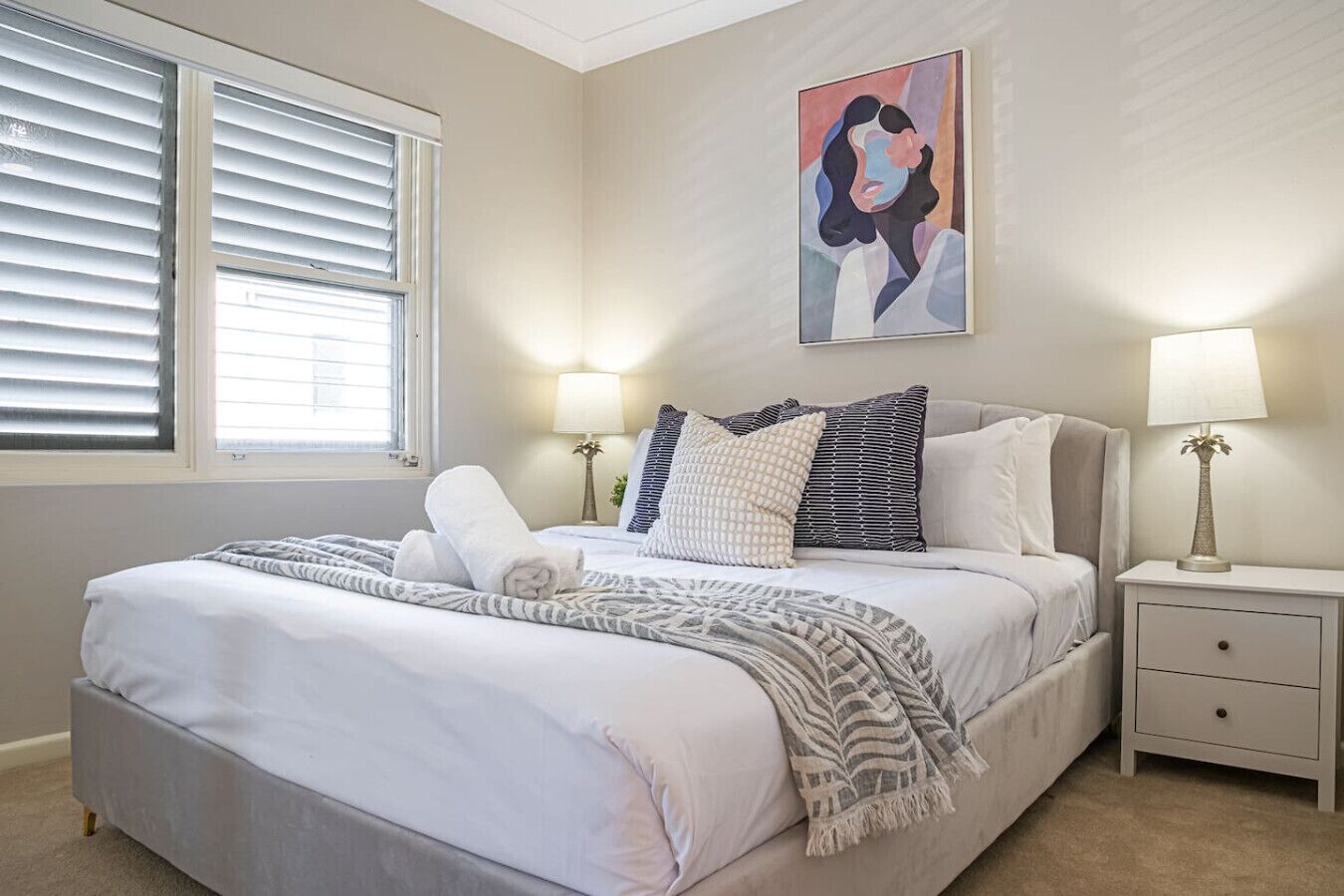 Een slaapkamer met een bed, twee nachtkastjes, een raam met jaloezieën en een schilderij aan de muur.
