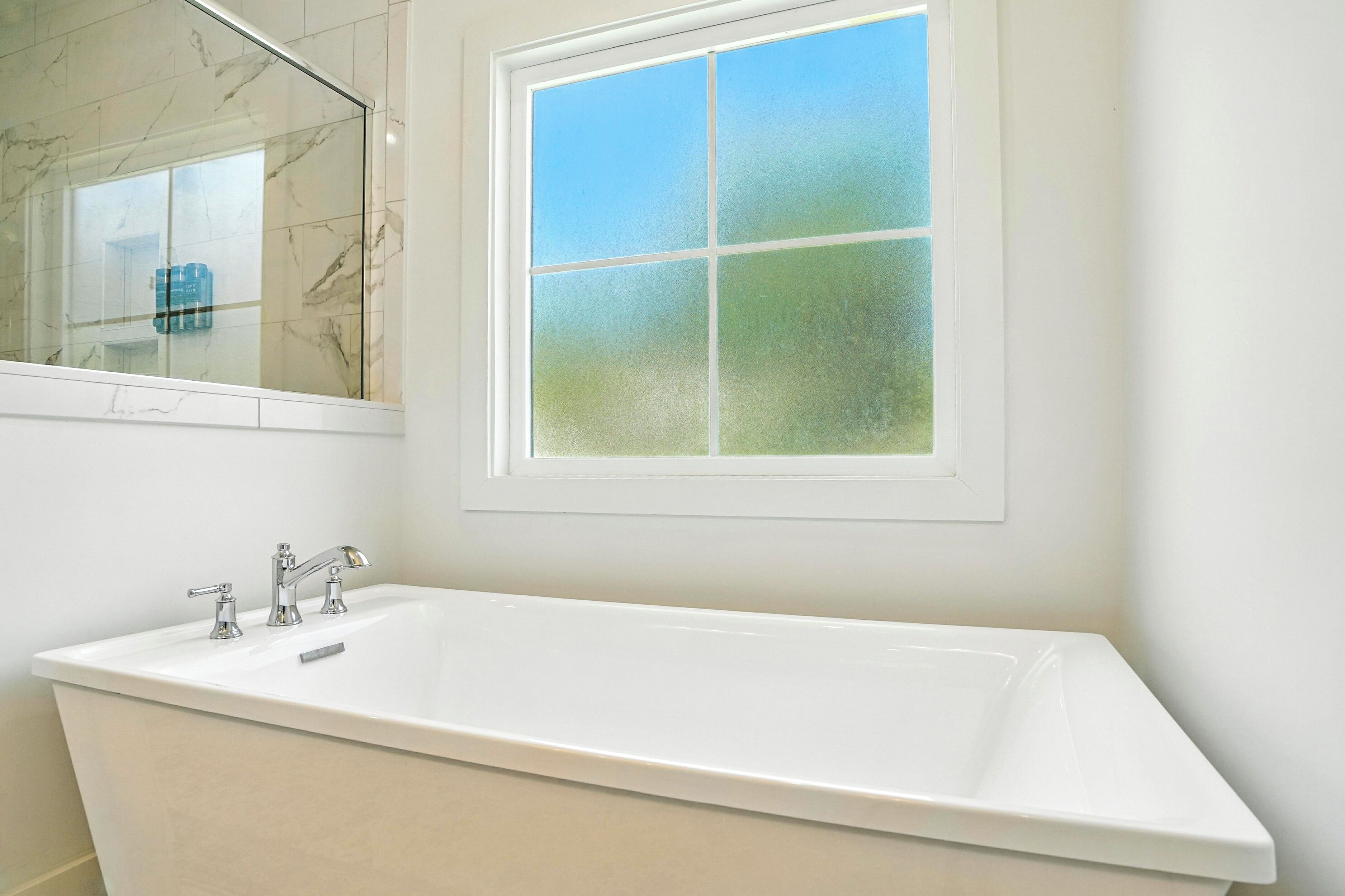 Un baño con una bañera blanca grande, un espejo con resalte de mármol y un ventanal esmerilado.