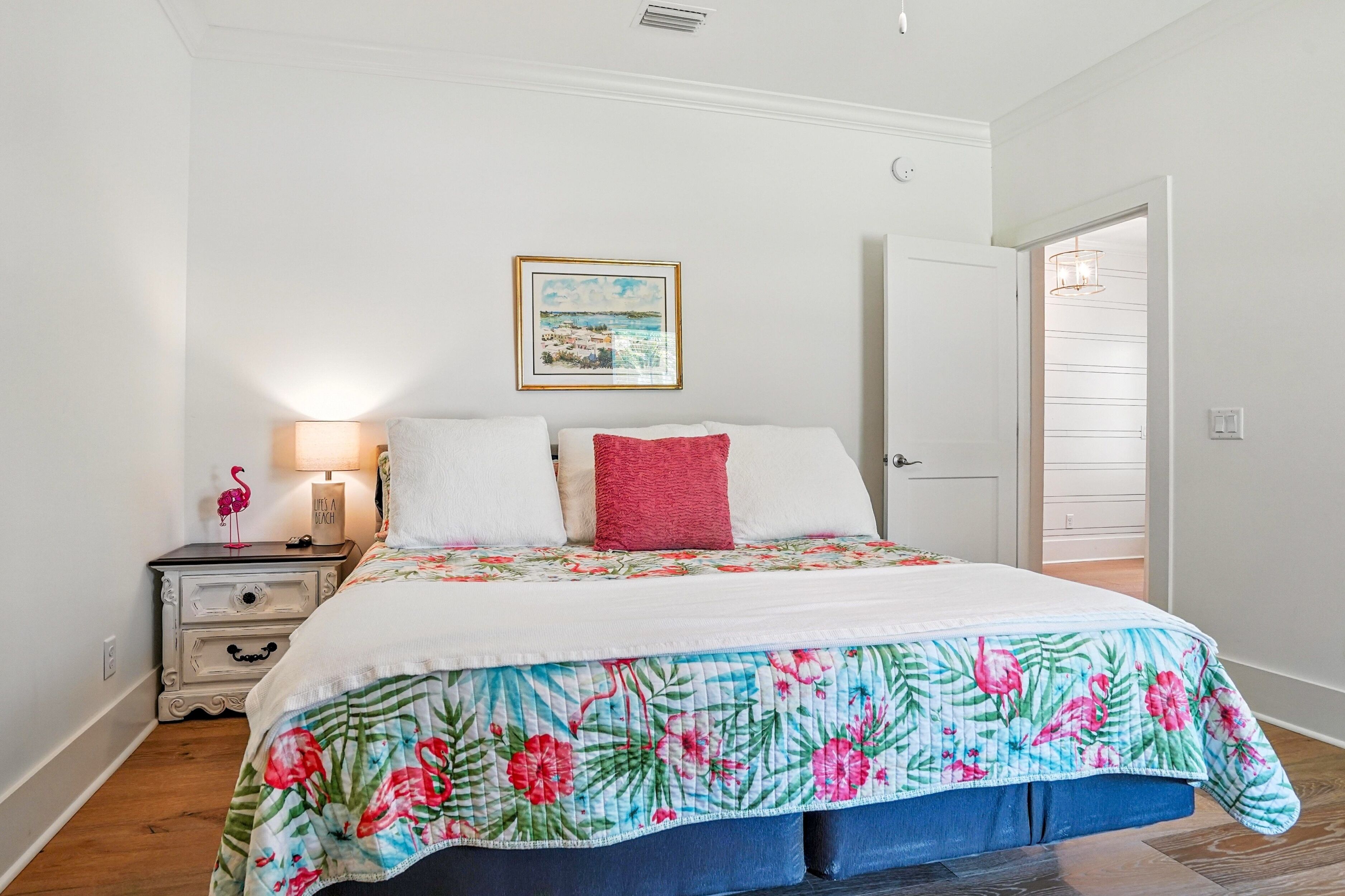A bedroom with a bed, a nightstand, a lamp, a door, and a wall with a framed picture.