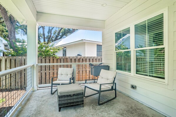 A covered porch with two chairs and a small table, a wooden fence, and a house in the background.