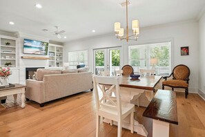 A spacious living room with a sofa, dining table, and chairs. There is a fireplace, a TV mounted on the wall, and a ceiling fan.