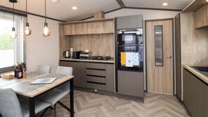 A modern kitchen with wooden cabinets, a built-in oven, and a microwave. There is a dining table with chairs and a window with blinds. - Hazelnut Lodge (Chipping Norton)