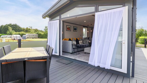 An open patio with a dining table and chairs, a sofa, and a sliding glass door leading to an interior space with a kitchen and seating area. - Hazelnut Lodge (Chipping Norton)