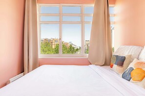 A bedroom with a large window, a bed with white linens, and a view of buildings and trees.