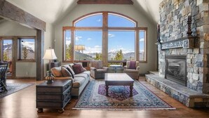 A spacious living room with a stone fireplace, large windows offering mountain views, a cozy seating area, and a wooden coffee table. - Top Of the World (Crested Butte)