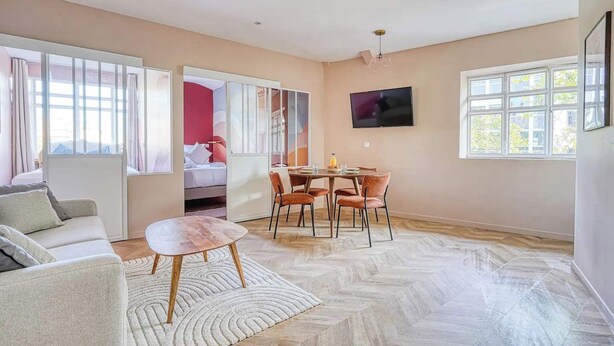 A modern living room with a sofa, a dining table with chairs, a TV mounted on the wall, and large windows providing natural light. - Climatisé & fonctionnel – 2BR cœur du 17ᵉ (Unknown)