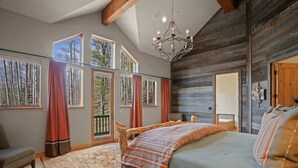 Quarto com cama grande, cabeceira de madeira, candelabro e janelas amplas com cortinas.
