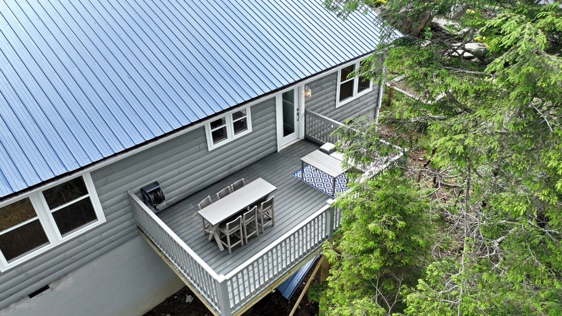 Ein zweistöckiges Haus mit blauem Metalldach, einem Balkon mit Tisch und Stühlen sowie einem Garten mit Bäumen.