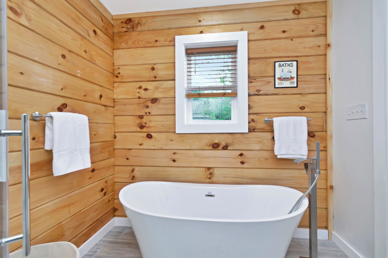 A wooden-paneled bathroom with a freestanding bathtub, two towel racks, and a window with a view of greenery.