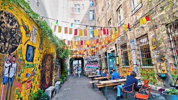 Un carrer estret amb taules i caires per a dinar a l'aire lliure, envoltat d'edificis de colors amb murals i grafitis.