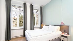 Un dormitori amb un llit, una tauleta de nit, una làmpada i una finestra amb cortines.