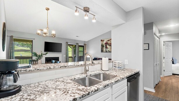 A modern kitchen with a granite countertop, stainless steel sink, and a dishwasher. There is a flat-screen TV mounted on the wall, a chandelier, and a window with a view of greenery.